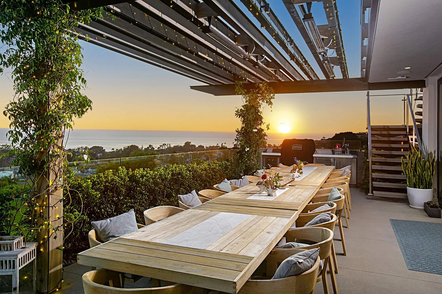 This image showcases a luxurious outdoor patio or deck area, perfect for entertaining. A long wooden dining table with comfortable chairs is the focal point, set against a stunning backdrop of the ocean at sunset. The space is covered by a modern pergola structure, adding both shade and architectural interest, while lush greenery and string lights enhance the ambiance.