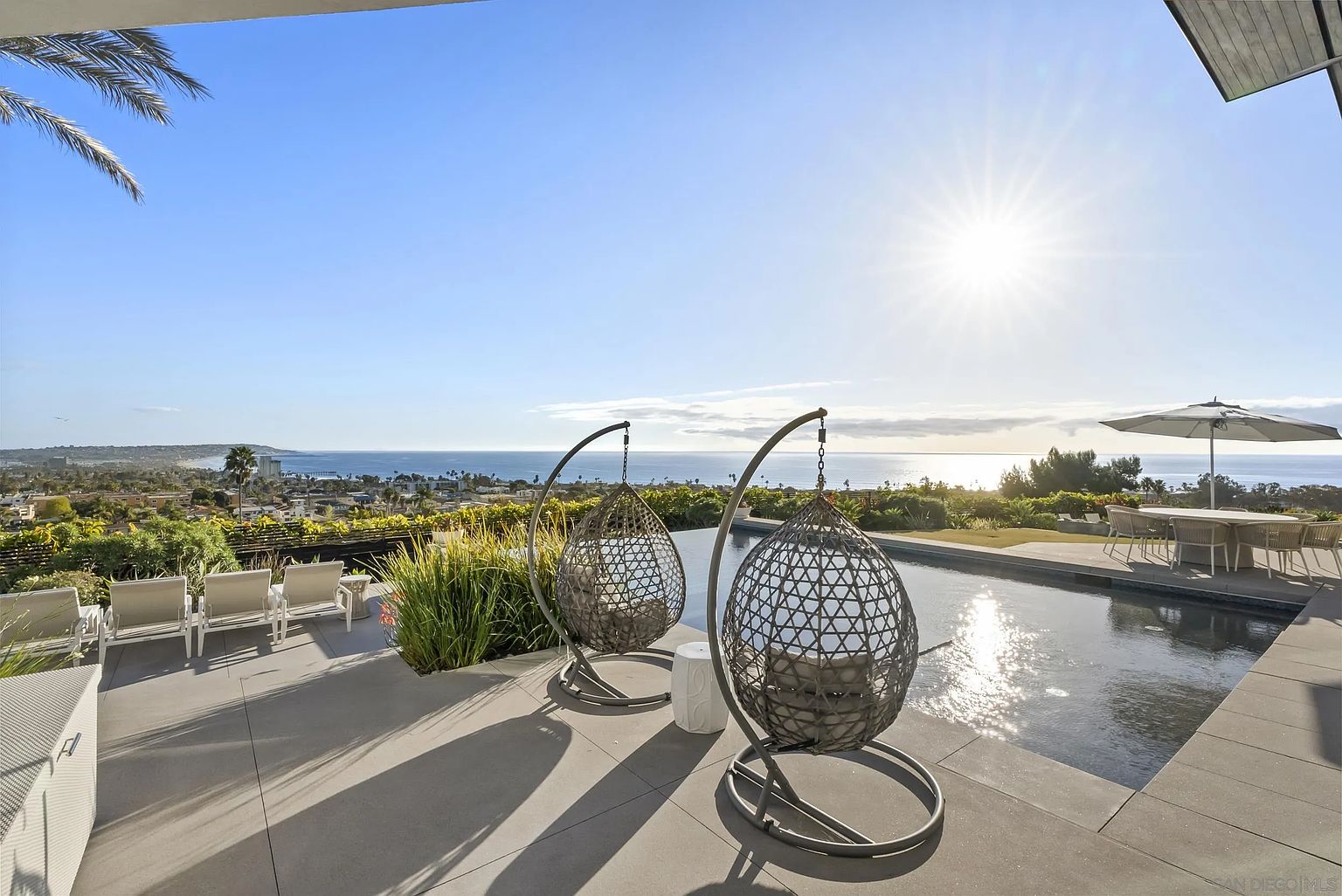 This image showcases a luxurious outdoor living space with a stunning pool overlooking the ocean. Two hanging chairs provide a relaxing spot to enjoy the view, while a dining set under an umbrella offers an area for outdoor meals. The sleek design and breathtaking scenery create an inviting and upscale atmosphere.