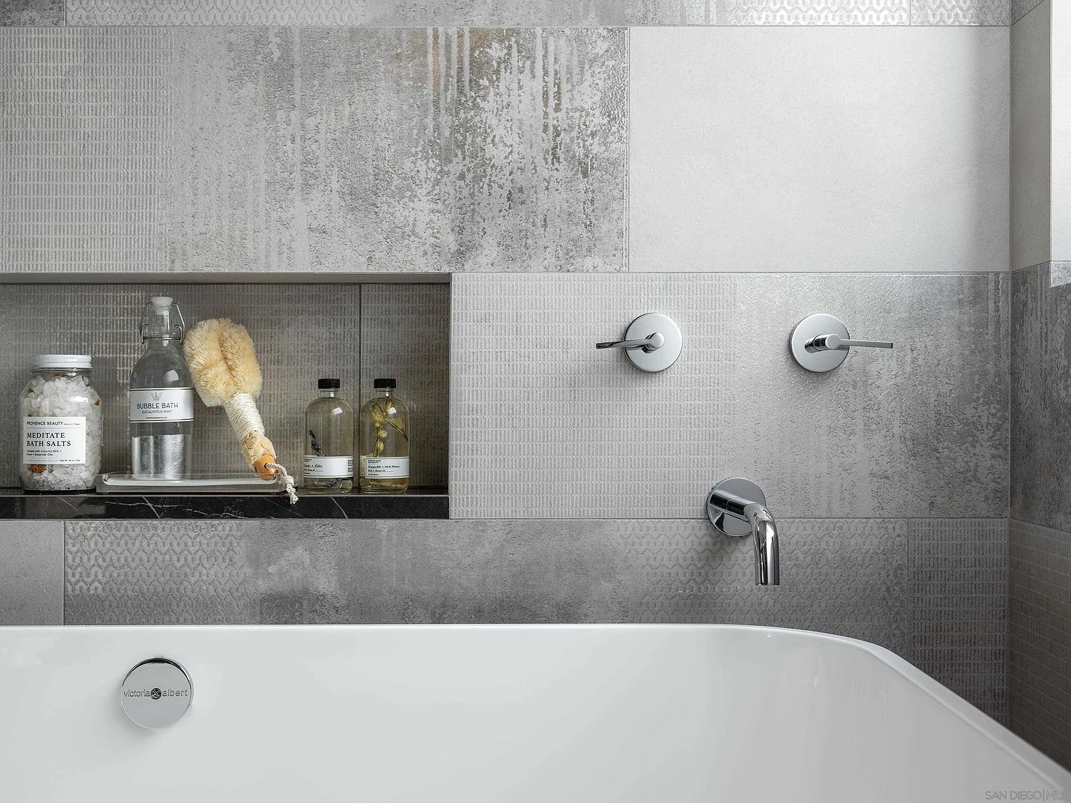 This image showcases a luxurious primary bathroom featuring a modern bathtub with chrome fixtures. The walls are adorned with a mix of textured and patterned gray tiles, creating a sophisticated and spa-like ambiance. A built-in shelf displays bath salts and other decorative items, adding to the room's serene aesthetic.