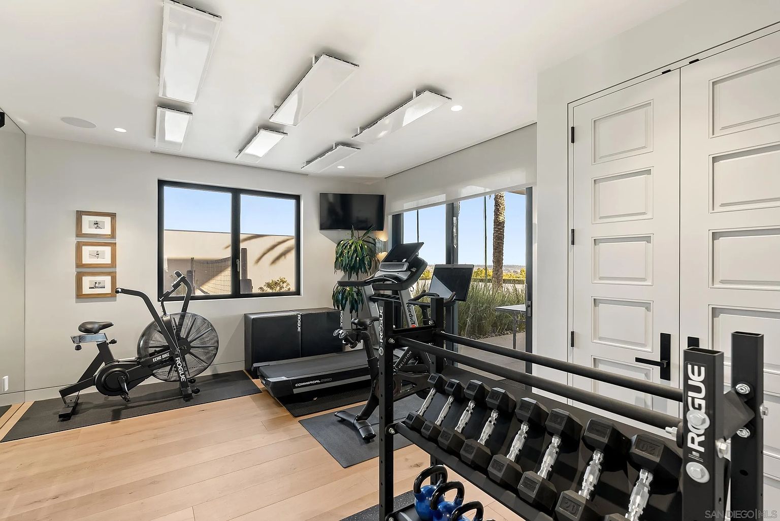 This is a well-equipped home gym featuring a variety of exercise equipment including a stationary bike, treadmill, and weight rack with dumbbells and kettlebells. The room has hardwood floors, large windows providing natural light, and a modern aesthetic with clean lines and neutral colors. The space appears organized and inviting for fitness activities.