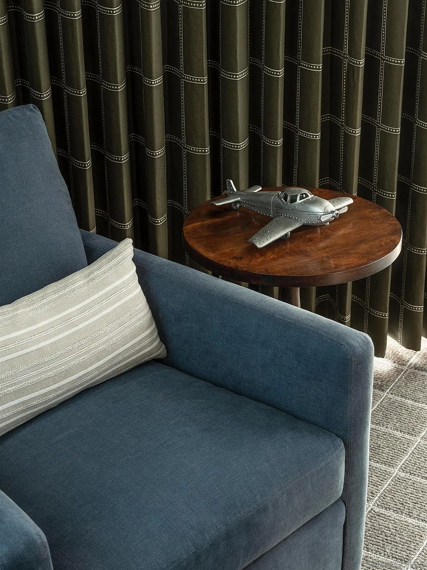 The image showcases a cozy living room setting with a blue armchair adorned with a striped pillow. A round wooden side table holds a decorative airplane model, adding a touch of character. The backdrop features patterned curtains, creating a warm and inviting atmosphere.