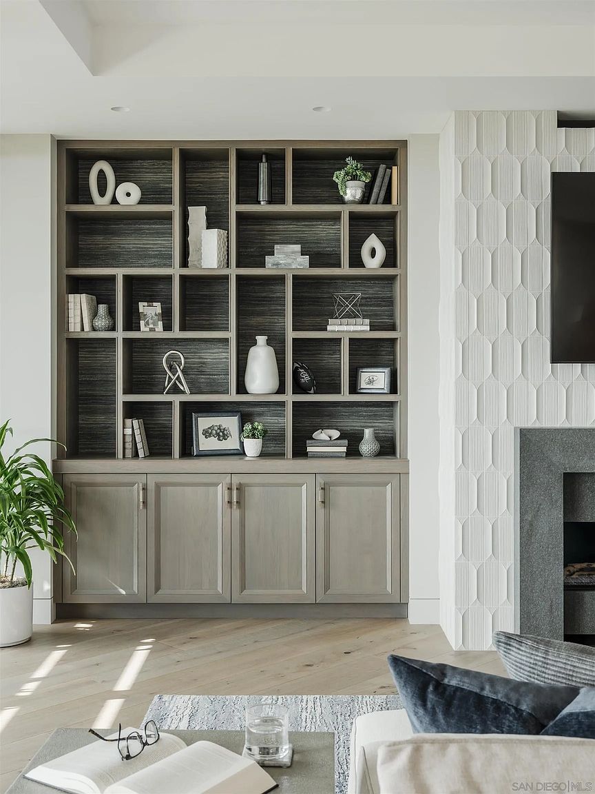 This image showcases a stylish living room featuring a built-in bookshelf with decorative items and closed storage below. The room has a modern aesthetic with a neutral color palette, complemented by a textured wall and hardwood flooring. The space is well-lit and appears inviting, suggesting a comfortable and sophisticated living area.
