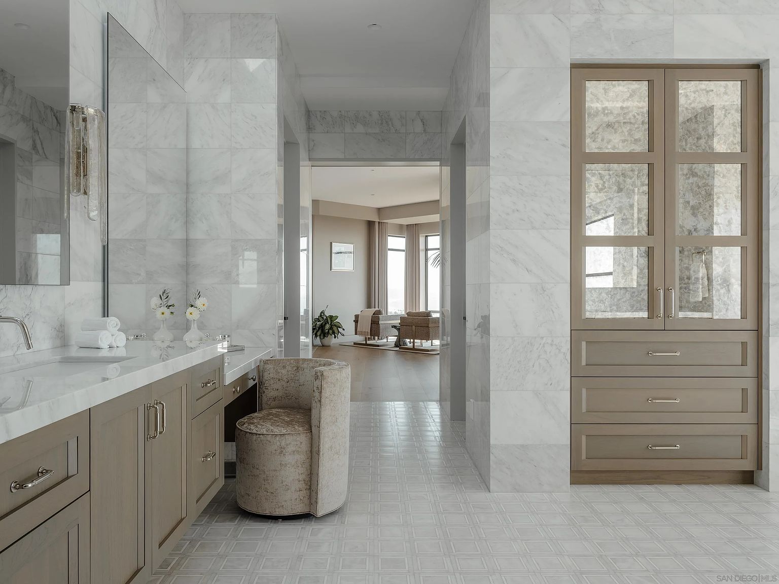 This is a luxurious primary bathroom featuring marble walls and floors, creating a bright and elegant space. The bathroom includes a vanity with light wood cabinets and a white countertop, a decorative chair, and a tall cabinet with glass doors. The perspective is from the doorway, looking into the bathroom and through to another room, giving a sense of spaciousness and high-end design.