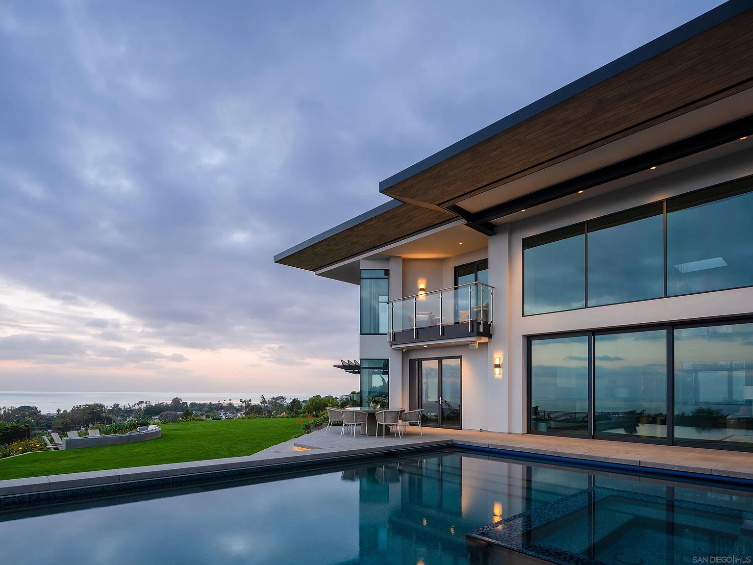This image showcases a modern luxury home with a stunning infinity pool overlooking the ocean. The architecture features clean lines, large glass windows, and a spacious balcony, blending indoor and outdoor living. The lush green lawn and serene pool create an inviting and upscale atmosphere.