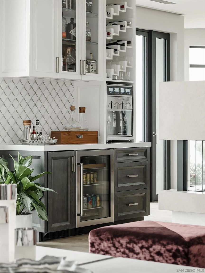 This image showcases a sophisticated wine cellar or bar area, featuring custom cabinetry with glass-fronted storage for spirits and a built-in wine refrigerator. The countertop is a light, solid surface, complemented by a diamond-patterned backsplash. The overall design exudes luxury and attention to detail, making it an attractive feature for potential buyers.