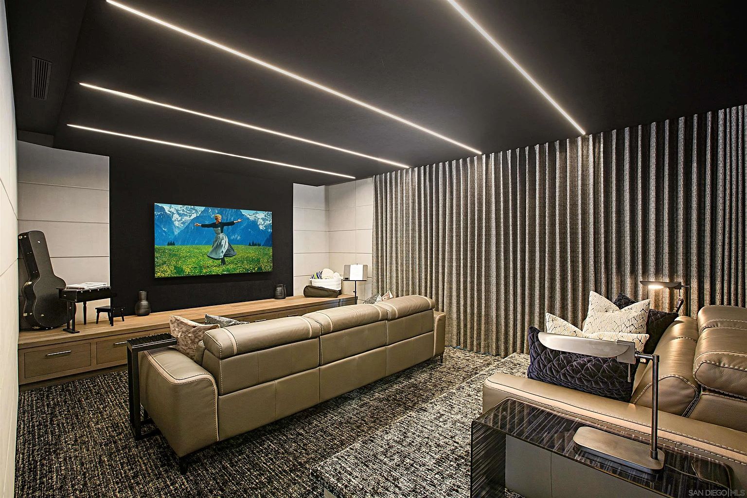 This is a modern living room featuring a large television displaying a nature scene, a comfortable-looking sectional sofa, and unique ceiling lighting. The room has a sophisticated and cozy ambiance, with a dark ceiling contrasting with lighter wall panels and a textured rug. The presence of a guitar and a small piano suggests a space for entertainment and relaxation.