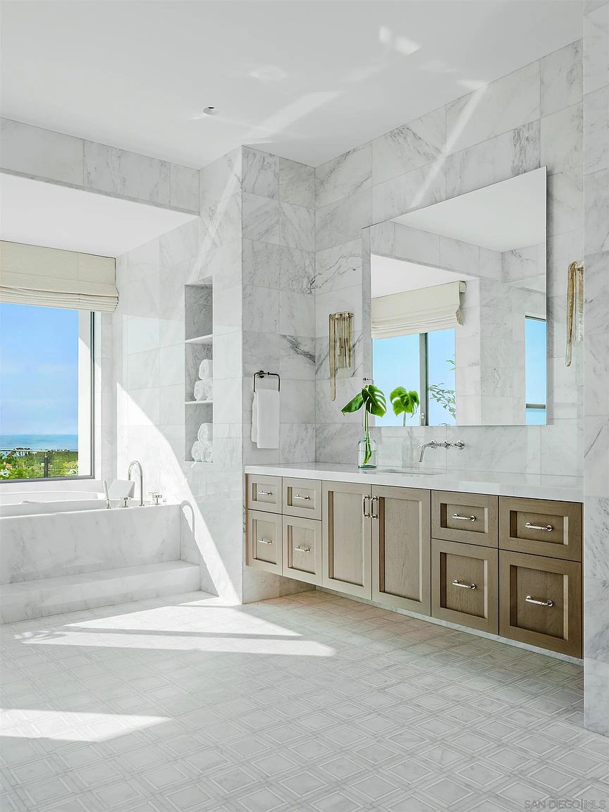This is a luxurious primary bathroom featuring marble walls and flooring. A large soaking tub sits beneath a window with ocean views, while a double vanity with custom cabinetry and a large mirror provides ample space. The room is bright and airy, creating a spa-like atmosphere.