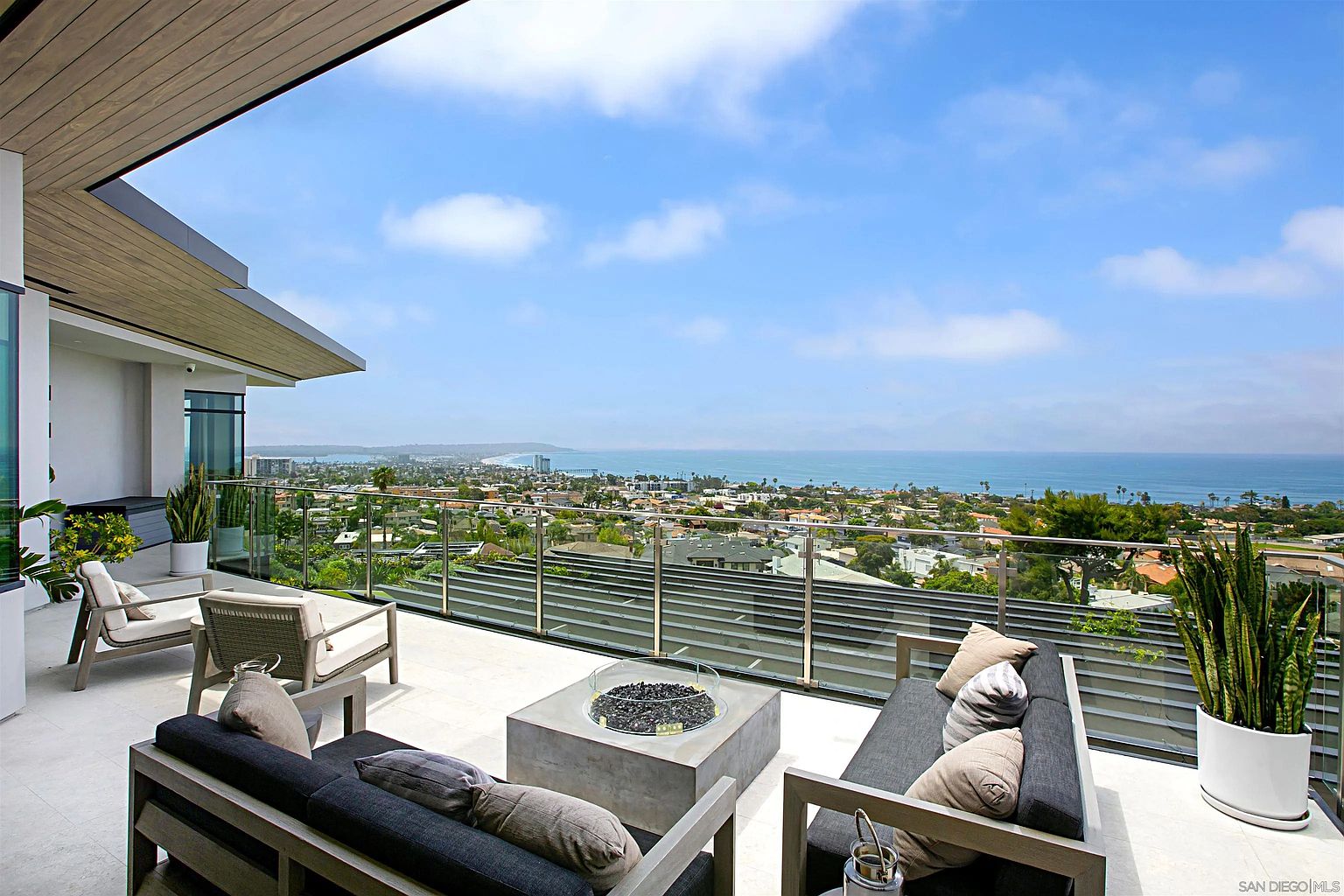 This image showcases a luxurious outdoor patio or balcony with stunning panoramic views of the city and ocean. The space is furnished with modern seating arrangements, including sofas and chairs with neutral-toned cushions, centered around a contemporary fire pit. The glass railing provides an unobstructed view, enhancing the appeal of this outdoor living space.