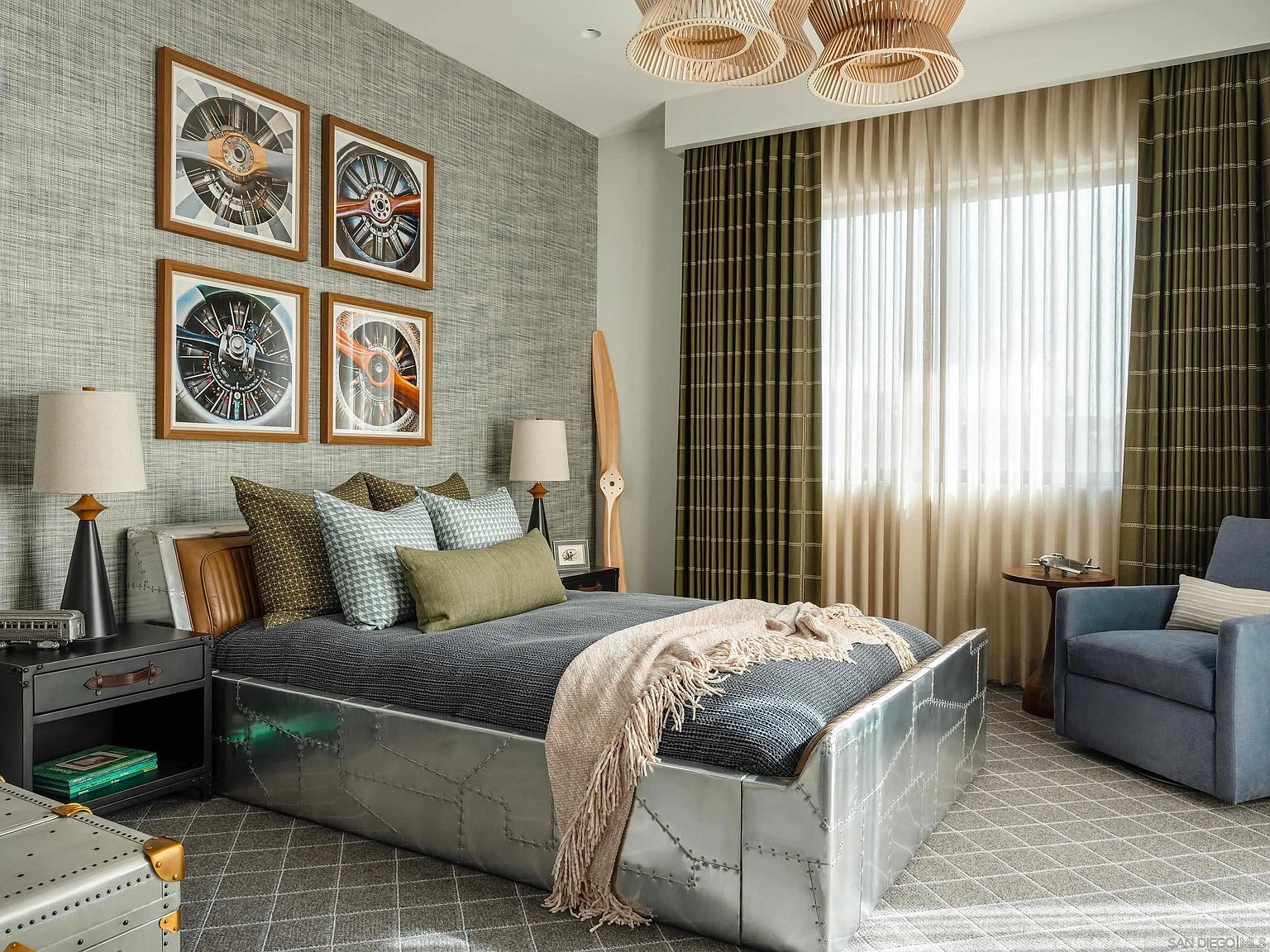 This is a stylish bedroom with an aviation theme, featuring a metal-clad bed frame, airplane propeller artwork, and a wooden propeller leaning against the wall. The room is decorated in neutral tones with pops of green and blue, creating a sophisticated and comfortable atmosphere. The window is covered with a combination of sheer and patterned curtains, allowing natural light to filter into the room.