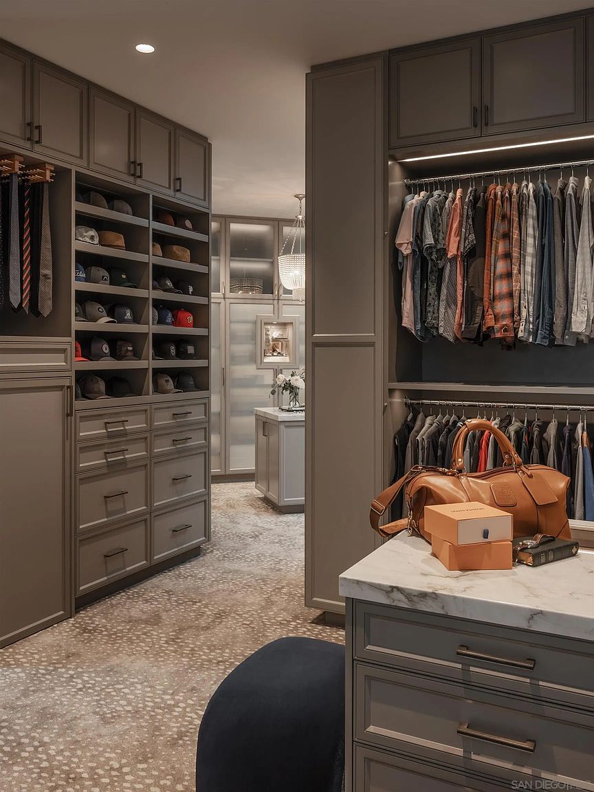 This is a well-organized walk-in closet featuring custom cabinetry in a neutral gray tone. The closet includes shelving for hats, drawers for clothing, and hanging rods for shirts and jackets. A marble-topped island provides a surface for accessories, and the overall design suggests a luxurious and functional space.
