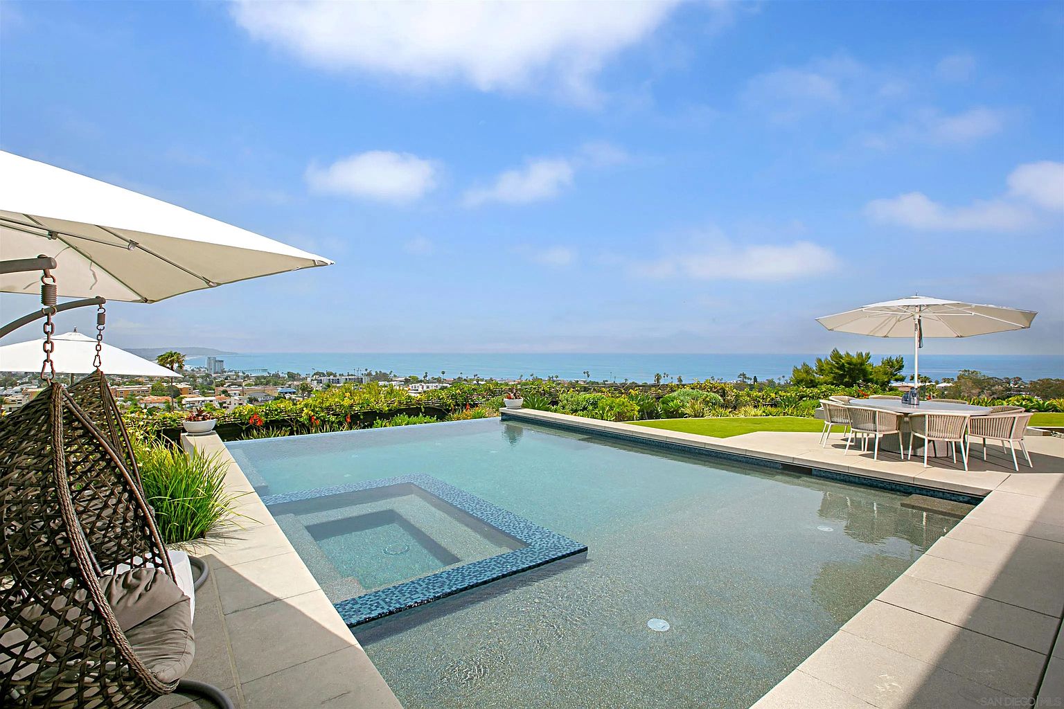This image showcases a luxurious outdoor pool and spa area with stunning ocean views. The pool features a built-in spa and is surrounded by a spacious patio with elegant outdoor furniture, including a dining set under an umbrella and a hanging chair. Lush landscaping adds to the serene and upscale ambiance, making it an ideal space for relaxation and entertainment.