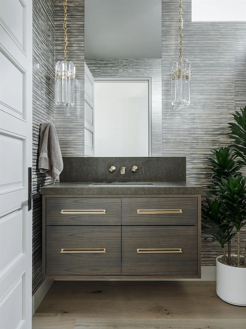 This is a stylish bathroom featuring a floating vanity with a dark wood finish and gold hardware. The walls are covered in a textured, horizontal-striped wallpaper, and the room is illuminated by modern pendant lights with crystal accents. A potted plant adds a touch of nature to the space, creating a sophisticated and inviting atmosphere.
