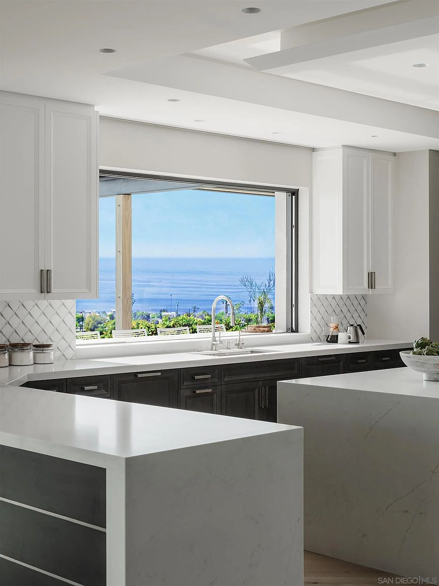 This is a bright and modern kitchen featuring white upper cabinets and dark lower cabinets, complemented by light countertops. A large window above the sink offers a stunning ocean view, enhancing the kitchen's appeal. The kitchen also includes a spacious island with a white countertop, adding to the functionality and style of the space.