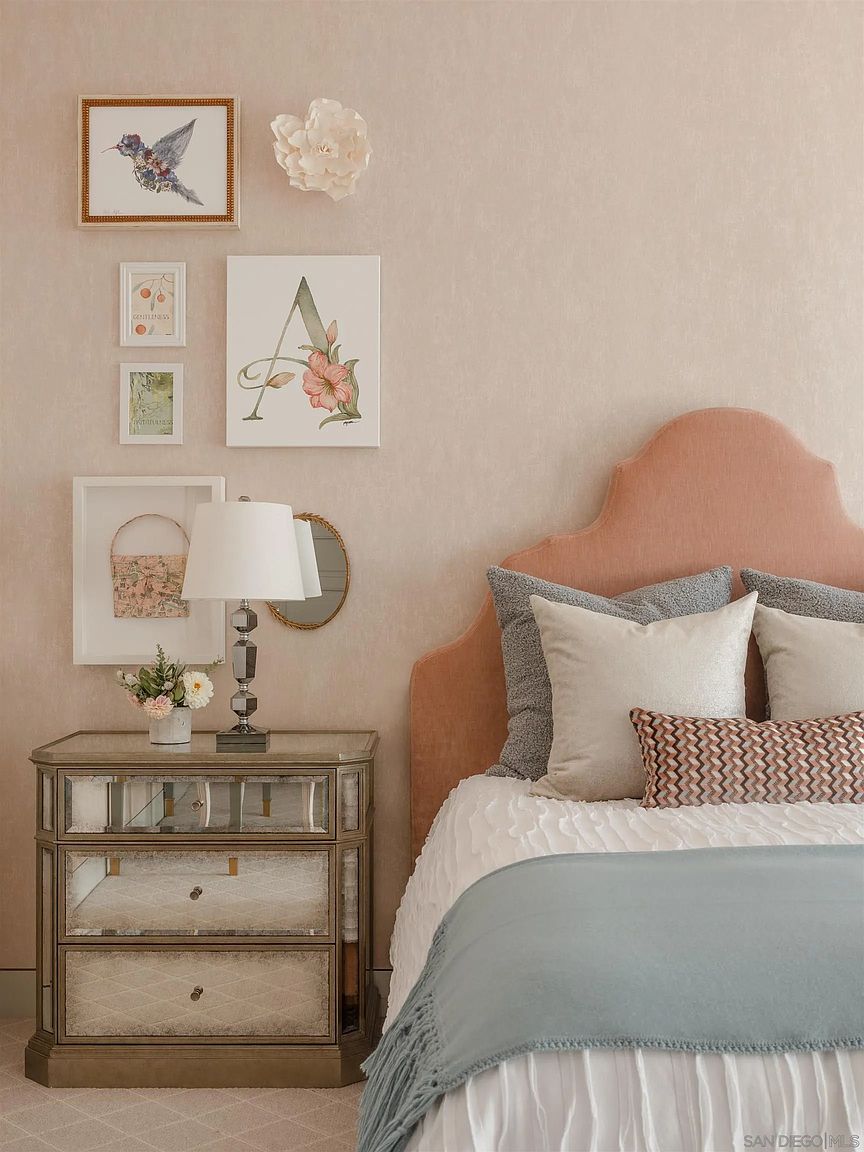 This is a charming bedroom featuring a pink upholstered headboard and a bed with white linens and a blue throw. A mirrored nightstand with a lamp sits beside the bed, adorned with decorative artwork and a small round mirror on the wall above. The room exudes a cozy and inviting atmosphere, perfect for a guest room.
