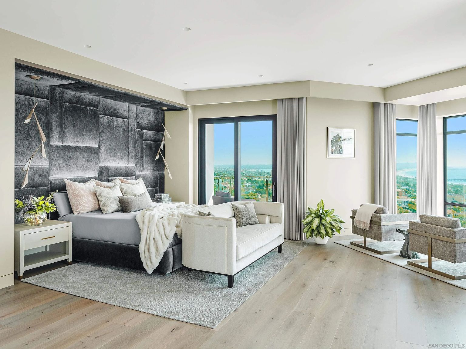 This is a luxurious primary bedroom featuring a large bed with a textured headboard and stylish lighting fixtures. The room includes a seating area with comfortable chairs and a sofa, complemented by large windows offering scenic views. The hardwood flooring and neutral color palette create a serene and sophisticated atmosphere.