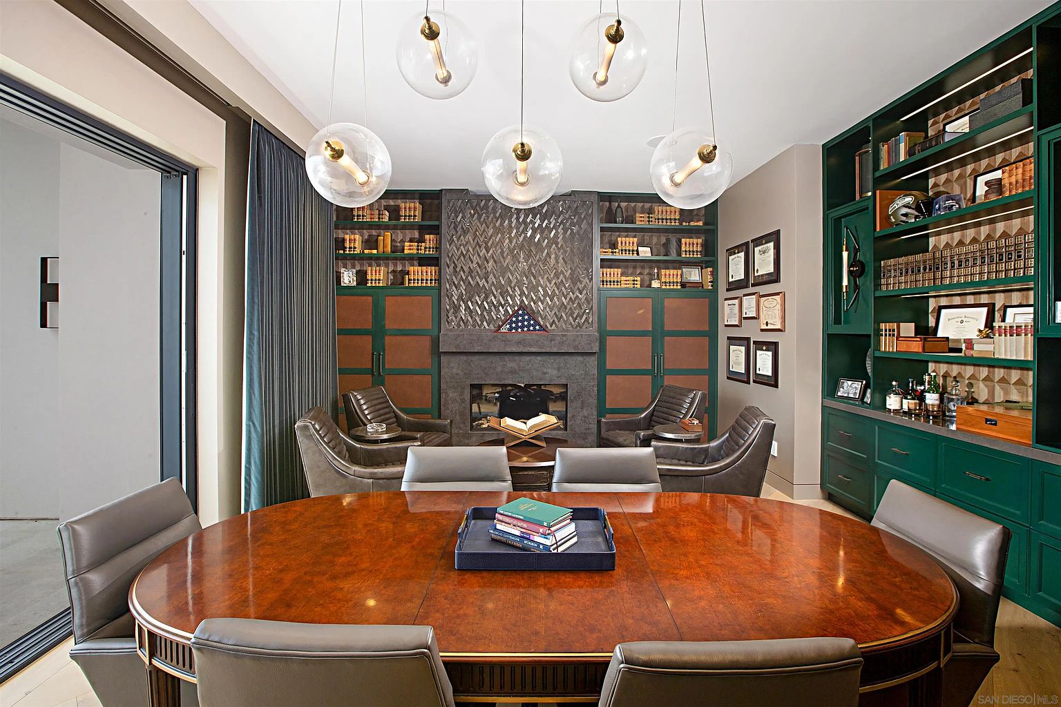 This is an interior shot of a sophisticated dining room featuring a large oval wooden table surrounded by leather chairs. The room is decorated with built-in bookshelves, a fireplace with a unique tile design, and modern globe pendant lights. The overall impression is one of elegance and refined taste, suitable for formal gatherings and intimate dinners.