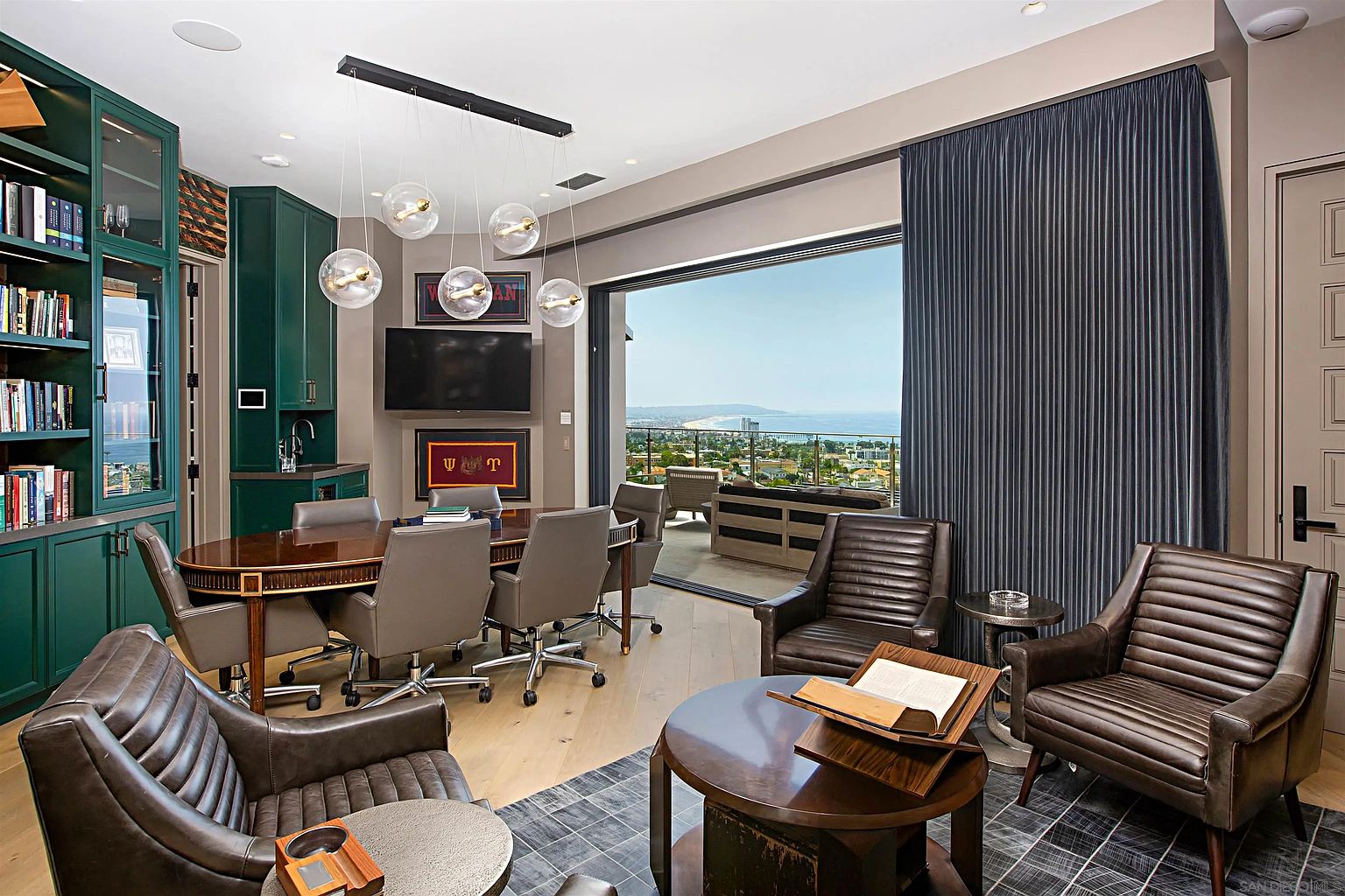 This is a well-appointed office or study featuring a large oval conference table surrounded by executive chairs, built-in shelving, and a wet bar. The room opens to an outdoor patio with a view of the ocean, creating a luxurious and productive workspace. The decor is sophisticated with dark wood accents and neutral tones.