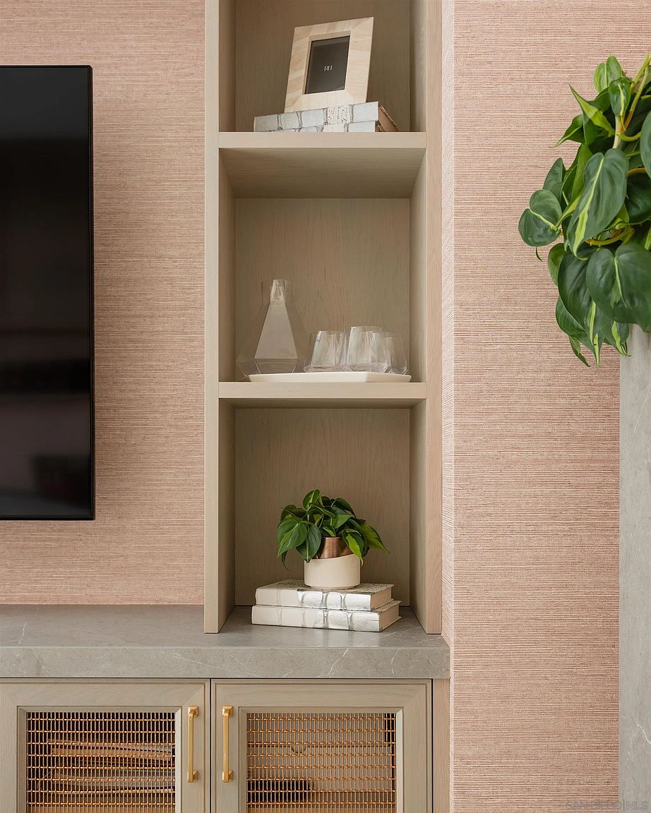 This image showcases a built-in shelving unit in a living room, featuring neutral-toned wallpaper and cabinetry with decorative mesh door fronts. The shelves are adorned with decorative items such as books, glassware, and plants, adding a touch of elegance and sophistication to the space. The overall impression is one of refined taste and attention to detail.