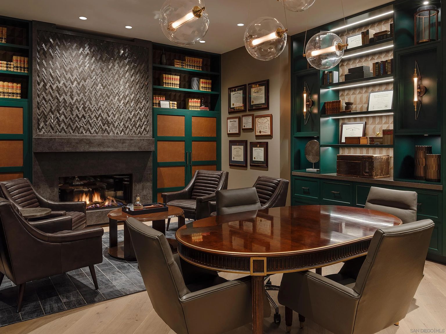 This is a sophisticated office or study featuring dark green built-in bookshelves filled with books and decorative items, complemented by a striking fireplace with a patterned tile surround. A large, oval-shaped wooden conference table is surrounded by leather chairs, creating a professional and inviting atmosphere. The room is illuminated by modern globe pendant lights, adding a touch of elegance.