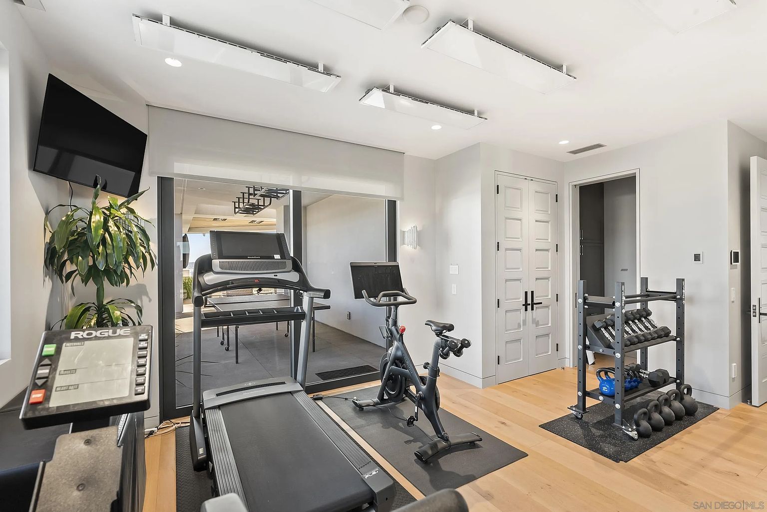 This is a well-equipped home gym featuring a treadmill, stationary bike, and weight rack with dumbbells and kettlebells. The room has light wood flooring, white walls, and modern lighting fixtures. A large sliding glass door provides natural light and access to an outdoor area, enhancing the workout environment.