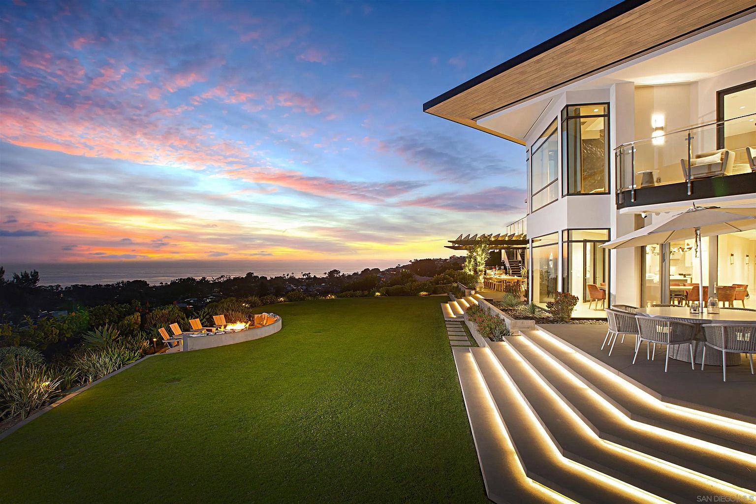 This image showcases the rear exterior of a modern luxury home at sunset. The property features a well-manicured lawn, a fire pit area with seating, and a multi-level patio with outdoor dining furniture. Illuminated steps lead from the patio to the lawn, and the home's architecture includes large windows and a balcony, offering stunning views of the ocean and surrounding landscape.