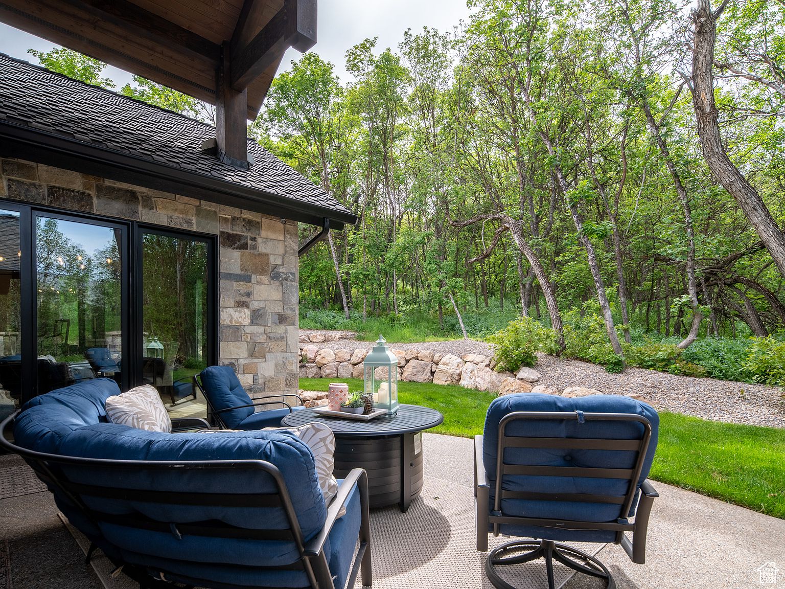 This image showcases a beautifully designed outdoor patio area, perfect for relaxation and entertaining. The patio features comfortable blue cushioned seating arranged around a modern fire pit table, creating a cozy ambiance. The space is surrounded by lush greenery and mature trees, offering privacy and a serene natural backdrop, while the stone facade of the house adds a touch of rustic elegance.