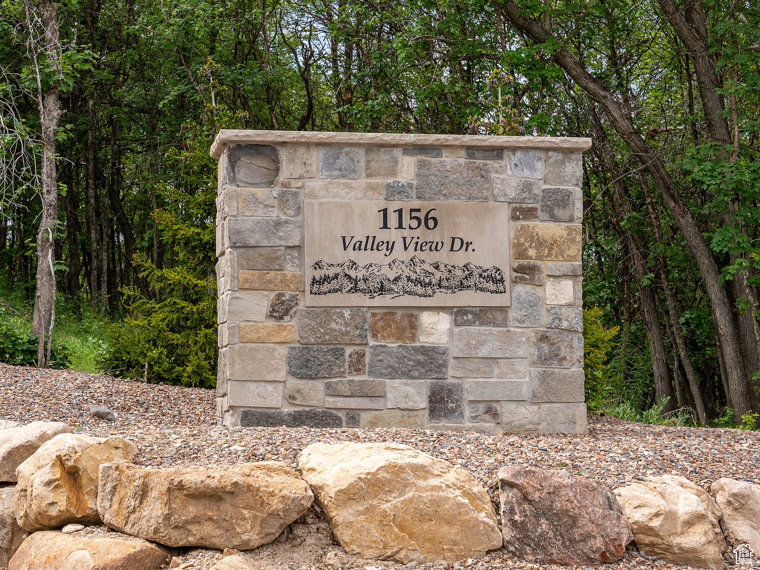 The image showcases a stone entrance sign for "1156 Valley View Dr." The sign is constructed from various shades of stone, creating a rustic yet elegant appearance. It is surrounded by gravel and large rocks, blending seamlessly with the natural landscape and suggesting a well-maintained property.