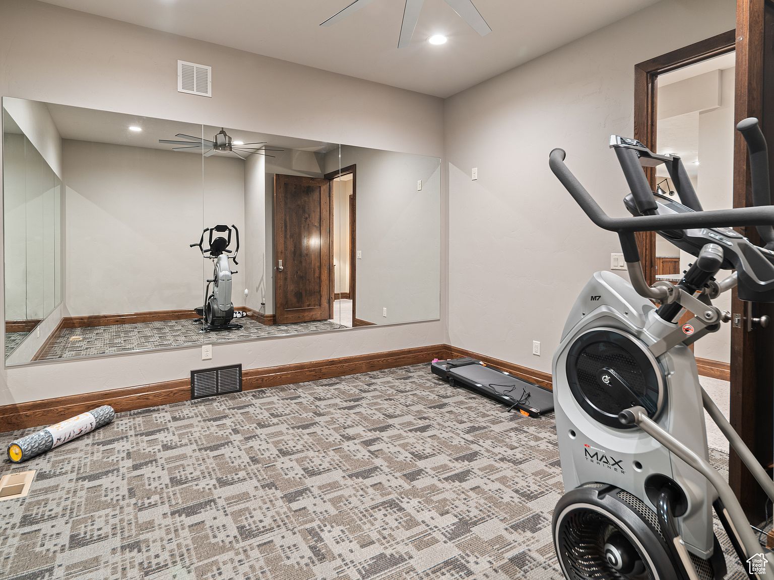 This is an interior shot of a home gym. The room features a large mirror, exercise equipment including an elliptical machine and a treadmill, and patterned carpeting. The walls are painted in a neutral tone, and the room has a clean, organized appearance, suggesting a dedicated space for fitness.