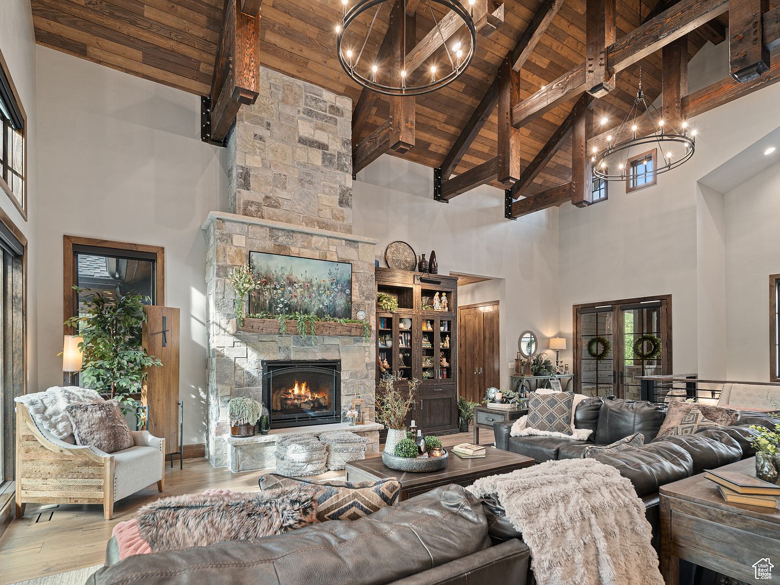This is a spacious living room featuring a stone fireplace with a mounted television, a large leather sectional sofa, and wooden ceiling beams with chandelier lighting. The room has a rustic yet luxurious feel, with a mix of textures and materials creating a warm and inviting atmosphere. The perspective is from the front of the room, capturing the full breadth of the space.