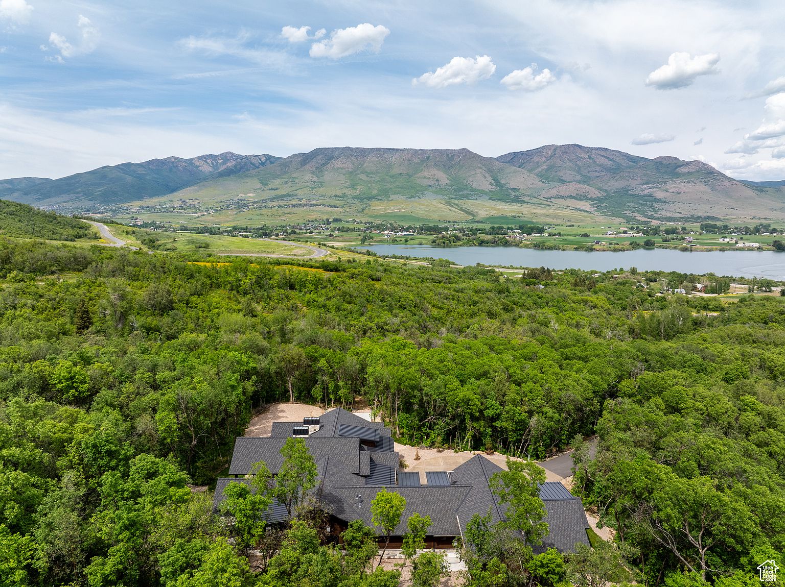 This aerial view showcases a luxurious home nestled amidst lush greenery, with a dark gray roof and a sprawling layout. The property is surrounded by dense trees, offering privacy and a sense of seclusion. In the background, rolling hills and a serene lake add to the picturesque setting, emphasizing the property's desirable location and stunning natural surroundings.