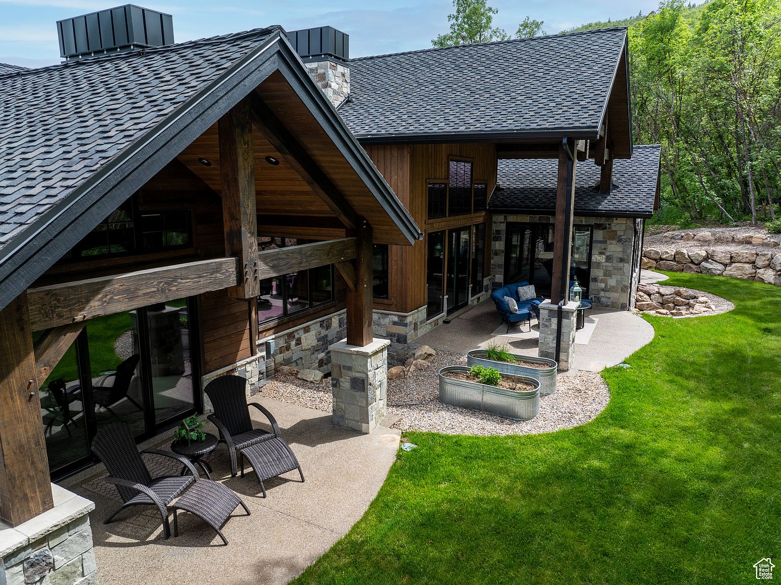 This is an aerial rear view of a luxurious home, showcasing its architectural design and outdoor living spaces. The house features a combination of wood and stone exterior, a dark shingled roof, and a spacious patio area with outdoor seating. The well-maintained lawn and landscaping enhance the property's appeal, creating a serene and inviting atmosphere.
