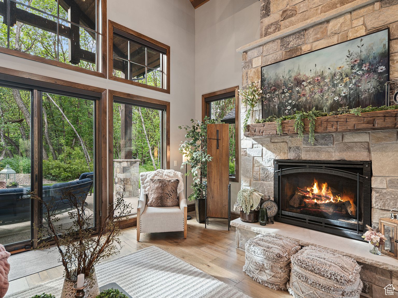 This inviting living room features a stone fireplace with a large painting above the mantel, creating a cozy focal point. Large windows and sliding glass doors offer views of the lush greenery outside, filling the space with natural light. The room is furnished with a comfortable armchair, decorative poufs, and a neutral-toned rug, creating a warm and sophisticated atmosphere.
