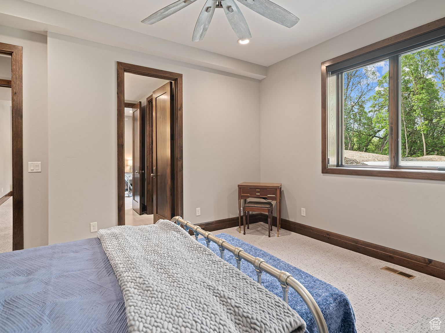 This is a well-lit bedroom featuring a metal-framed bed with blue and gray bedding. The room has neutral-colored walls, carpeted floors with dark wood trim, and a window offering a view of trees outside. A small wooden desk sits against the wall, and a doorway leads to another room, creating a sense of spaciousness.