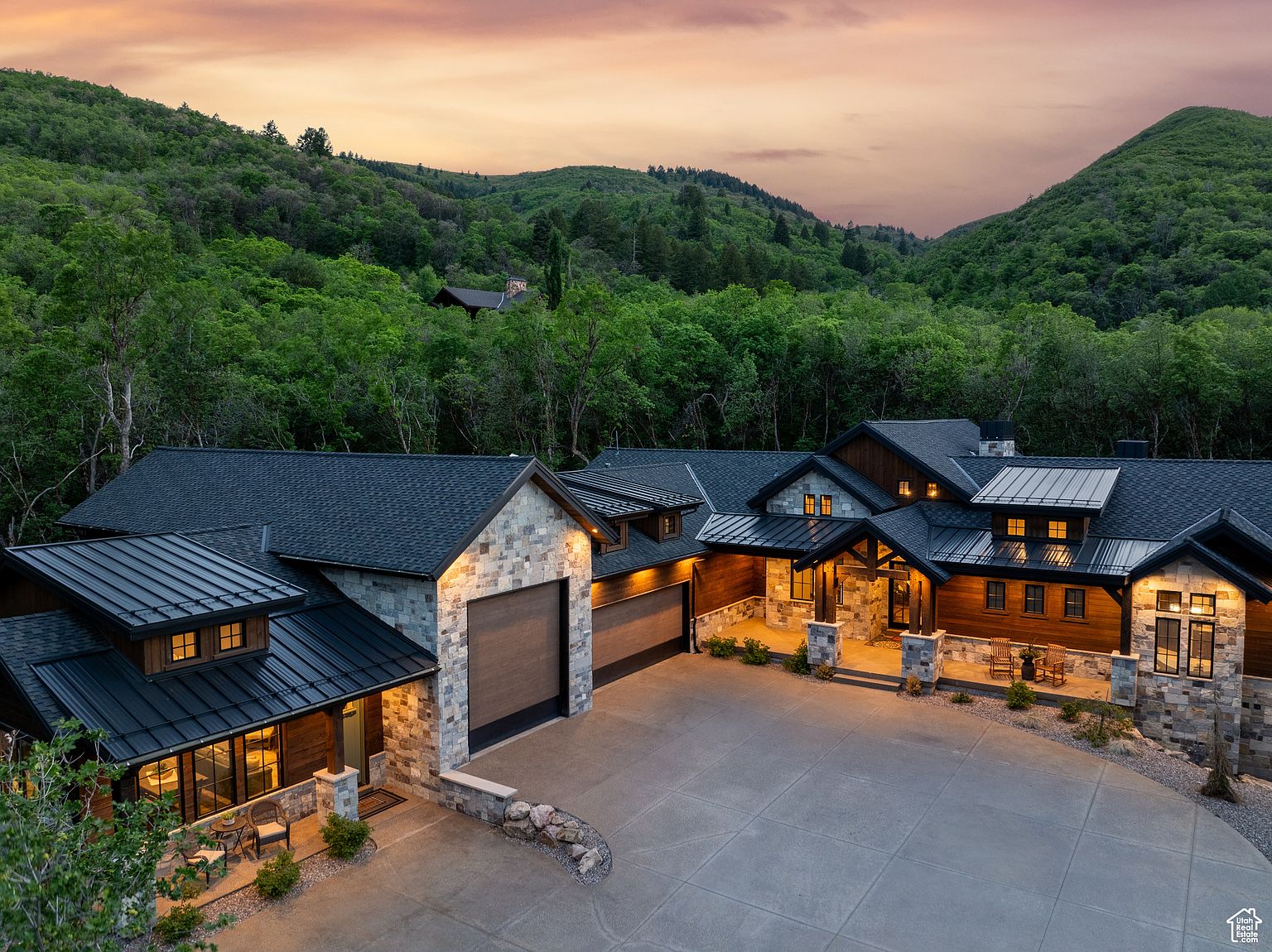 This aerial view showcases a luxurious home nestled in a lush, green valley. The house features a combination of stone and wood exterior, dark metal roofing, and multiple garage bays. A spacious concrete driveway leads to the entrance, and the surrounding landscape provides a sense of privacy and tranquility.