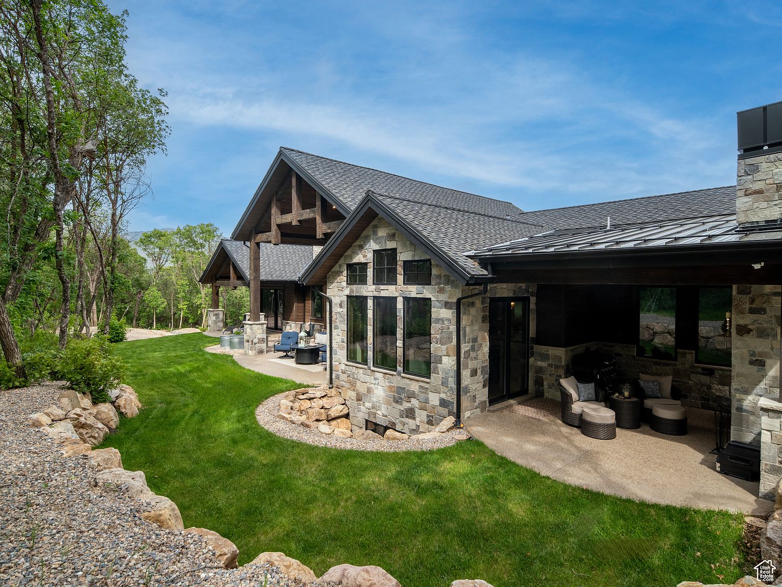 This image showcases the rear exterior of a luxurious home, emphasizing its stone facade, dark-framed windows, and covered patio area with comfortable seating. The well-manicured lawn and landscaped rock features add to the property's appeal, creating an inviting outdoor living space. The architectural design blends natural materials with modern elements, offering a sophisticated and serene atmosphere.