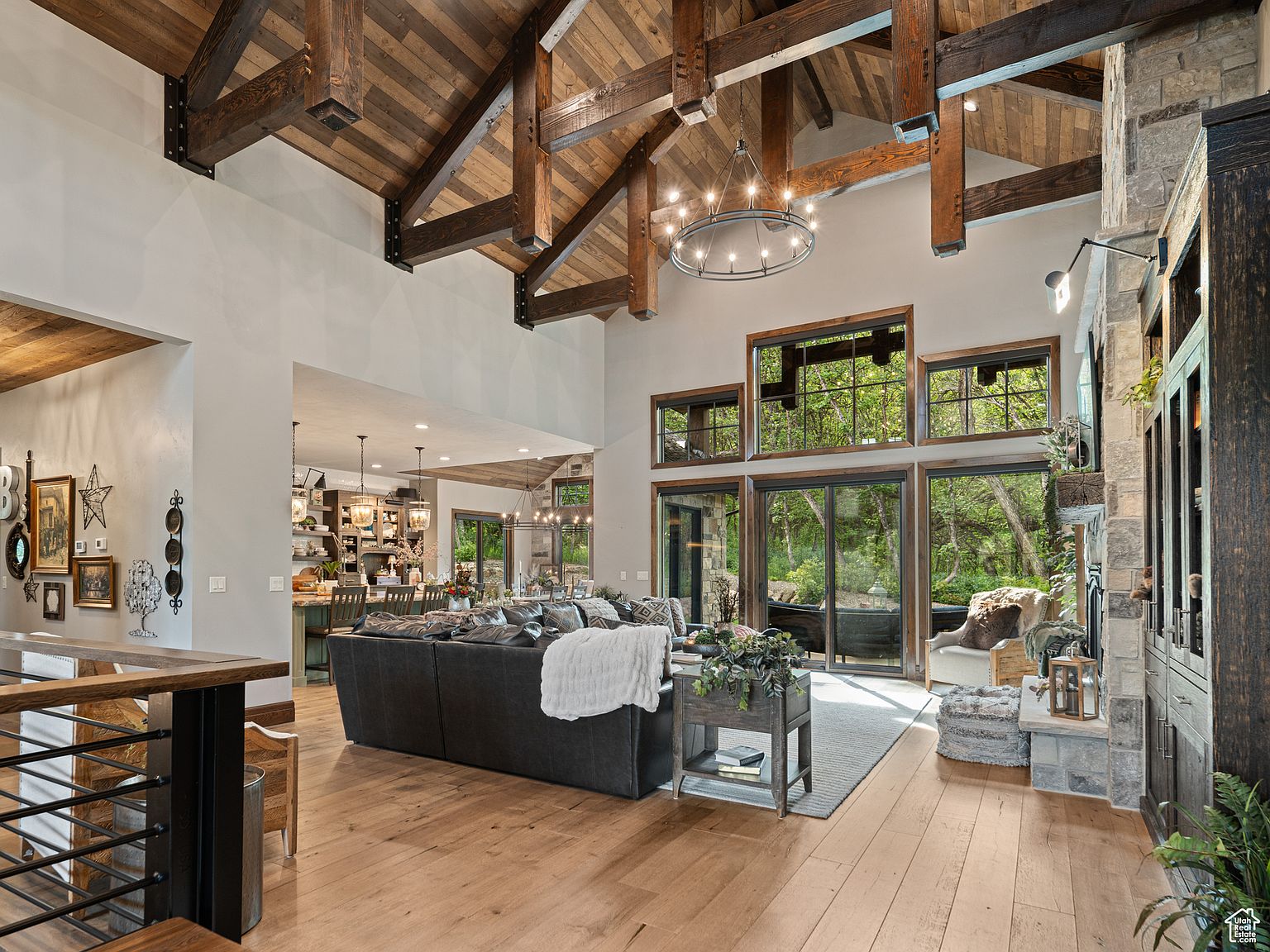 This is a spacious living room featuring a high vaulted ceiling with exposed wooden beams, creating a rustic yet elegant atmosphere. Large windows offer views of the surrounding greenery, filling the room with natural light. The room is furnished with a comfortable sectional sofa, a stone fireplace, and tasteful decor, making it an inviting space for relaxation and entertainment.