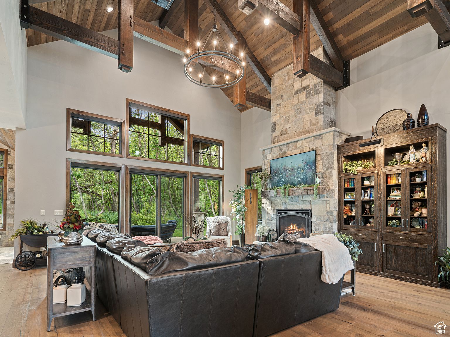 This is an interior shot of a luxurious living room featuring a high vaulted ceiling with exposed wooden beams and a stone fireplace. Large windows offer a view of the lush greenery outside, and a comfortable leather sectional sofa anchors the space. A large chandelier hangs from the ceiling, adding to the room's elegant ambiance.