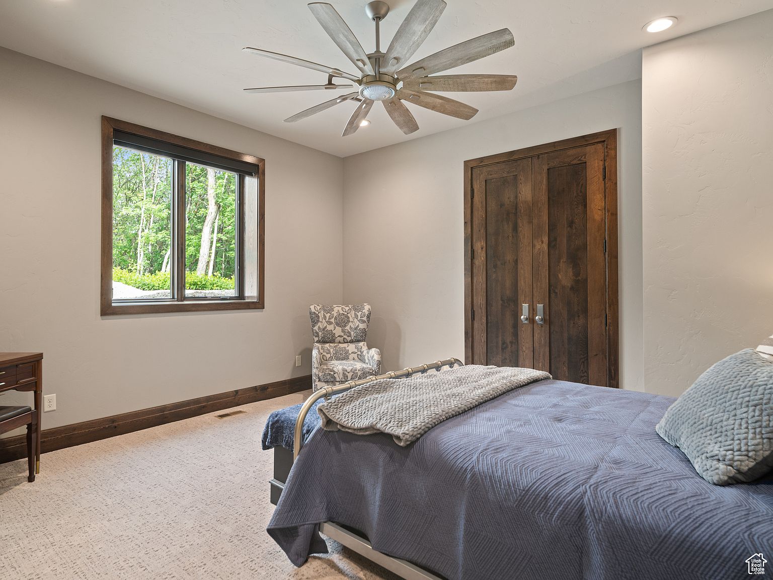 This is a cozy guest bedroom featuring a bed with a blue quilt and a gray throw blanket. The room has a large window providing natural light and a view of the outdoors, complemented by a decorative chair and a wooden-framed closet. A ceiling fan adds to the room's comfort, and the overall impression is inviting and serene.
