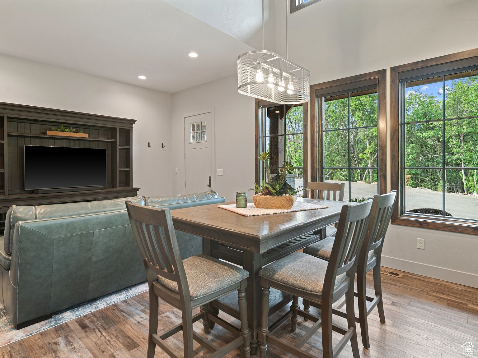 This interior shot showcases a dining area adjacent to a living space, featuring a wooden dining table with four chairs and a modern chandelier. Large windows provide ample natural light and views of the surrounding greenery. The room exudes a cozy and inviting atmosphere, perfect for family meals and gatherings.