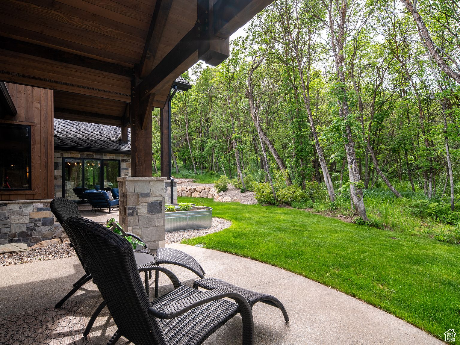 This image showcases a beautifully designed outdoor patio area. The patio features comfortable wicker lounge chairs, a concrete surface, and a view of a lush green lawn and wooded area. The architectural details of the home, including stone accents and wooden beams, add to the overall appeal of this outdoor living space.