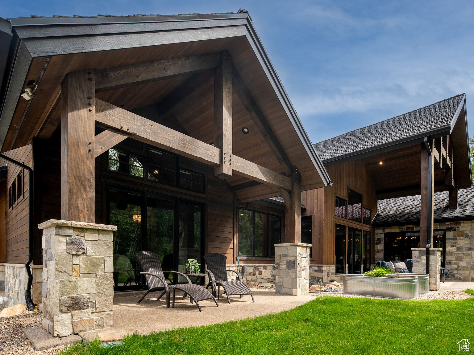This image showcases a beautifully designed patio area of a modern home. The patio features comfortable lounge chairs, stone pillars supporting a wooden structure, and a well-maintained lawn. The architectural details, such as the dark wood and stone accents, create a luxurious and inviting outdoor space.