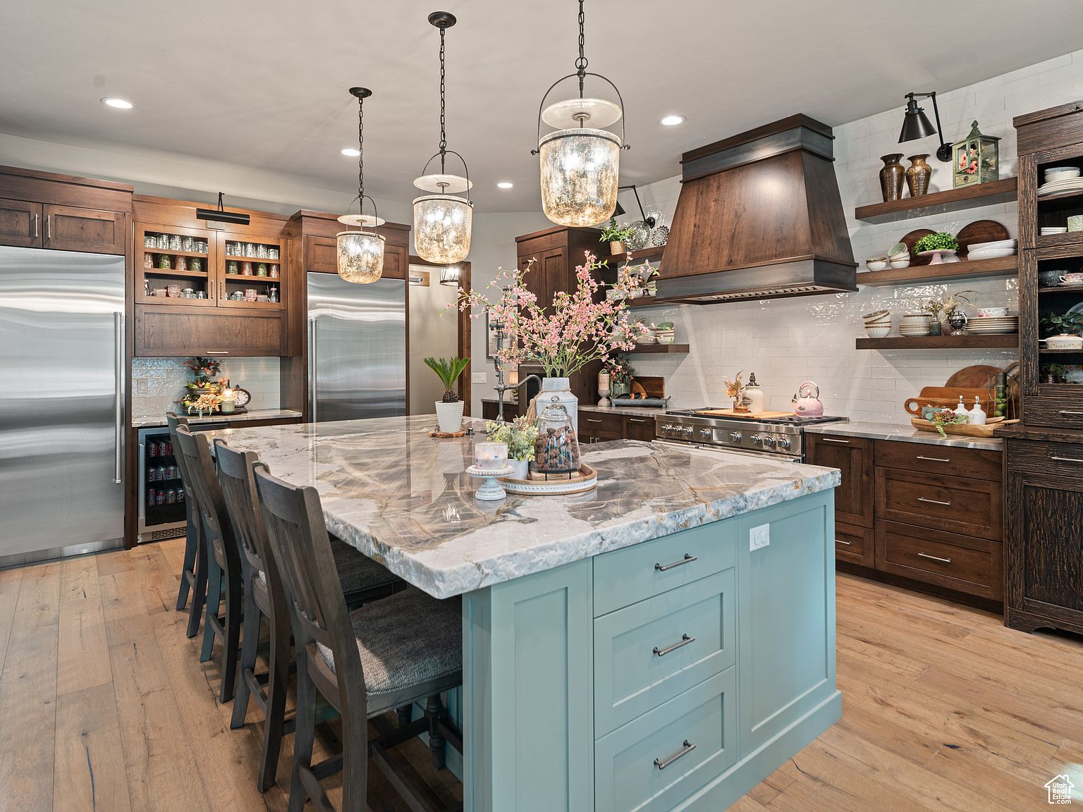 This is a spacious kitchen featuring a large island with a unique countertop and light blue cabinetry. The kitchen is equipped with stainless steel appliances and dark wood cabinets, complemented by open shelving displaying decorative items. Three pendant lights hang above the island, adding to the warm and inviting atmosphere.