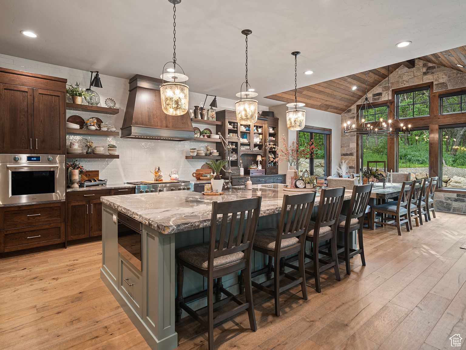 This is a spacious kitchen featuring a large island with seating, custom cabinetry, and high-end appliances. The kitchen seamlessly transitions into a dining area with large windows offering views of the outdoors. The overall design combines rustic and modern elements, creating a warm and inviting atmosphere.