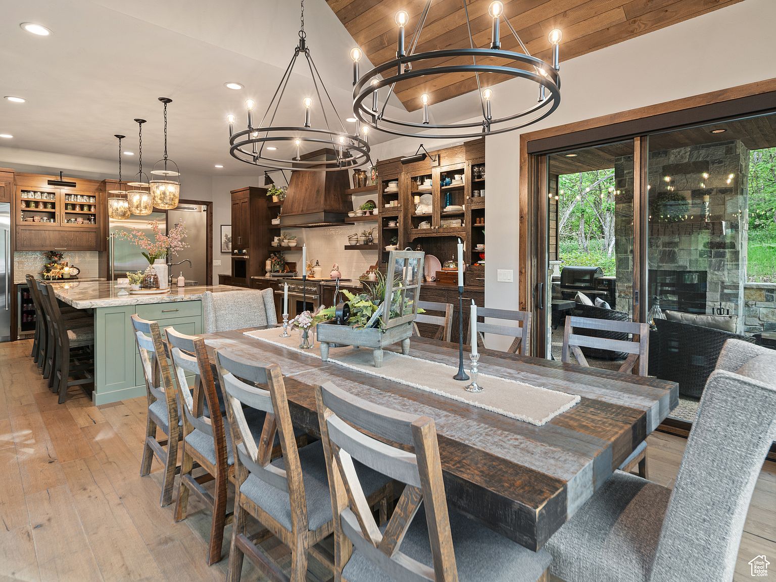 This interior shot showcases a dining room with a large, rustic wooden table surrounded by chairs with gray cushions. Above the table hang two circular chandeliers, adding a modern touch to the rustic aesthetic. The dining area seamlessly connects to the kitchen and offers a view of an outdoor patio through sliding glass doors, creating an inviting and open space.