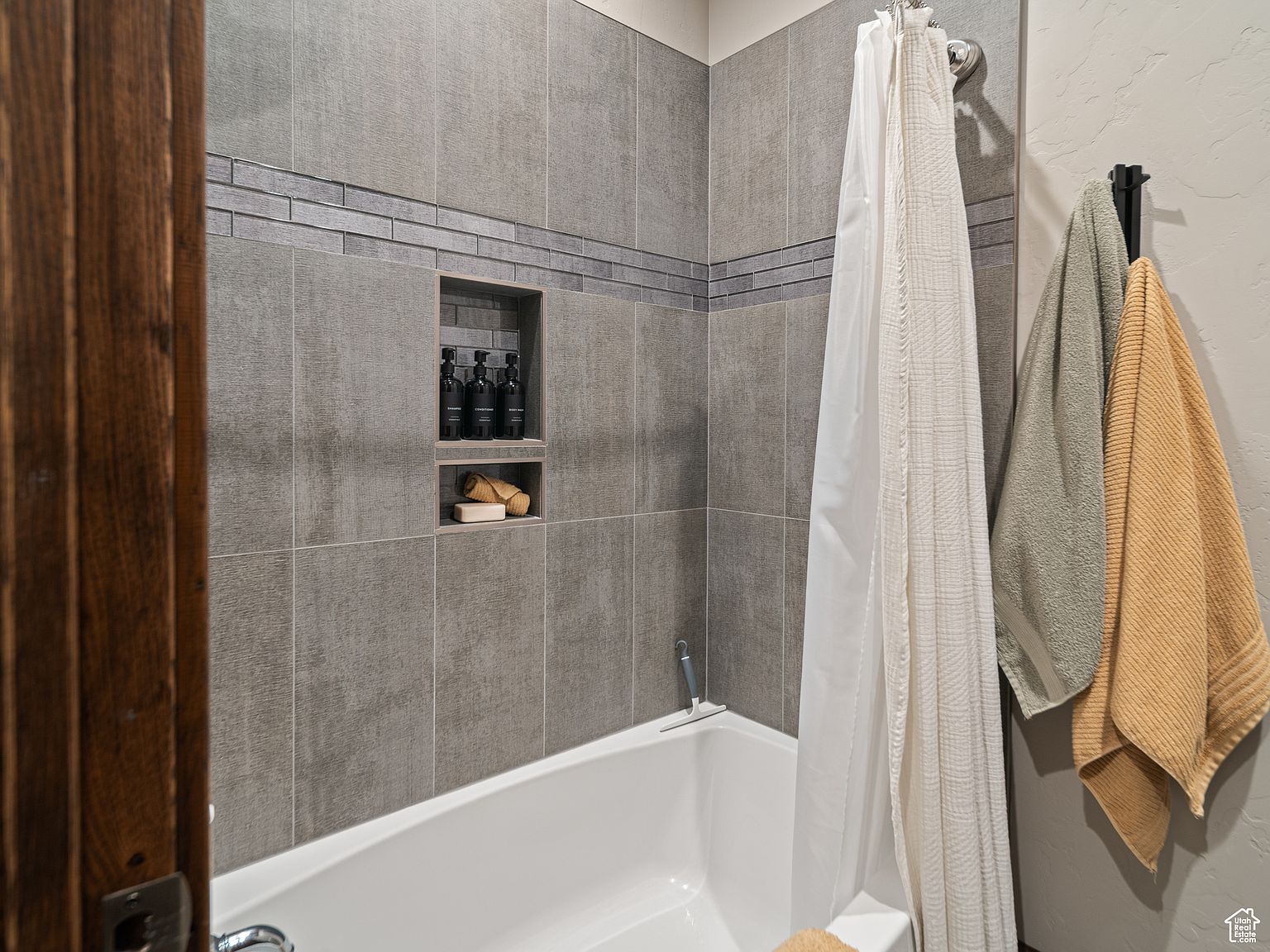 This is a well-appointed bathroom featuring gray tiled walls with a decorative accent strip and a built-in niche for toiletries. A white bathtub is partially visible, and a shower curtain hangs to the side. Two towels are neatly hung on a black rack, adding a touch of color and texture to the space.