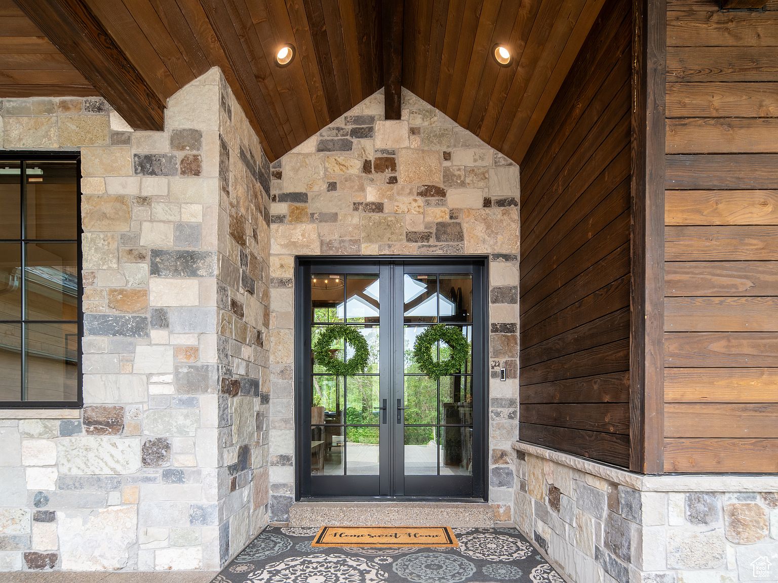 This image showcases a grand entryway featuring a double door with black frames and decorative wreaths. The exterior is constructed with a mix of stone and wood, creating a rustic yet elegant aesthetic. A patterned rug welcomes visitors, enhancing the inviting atmosphere of the home.