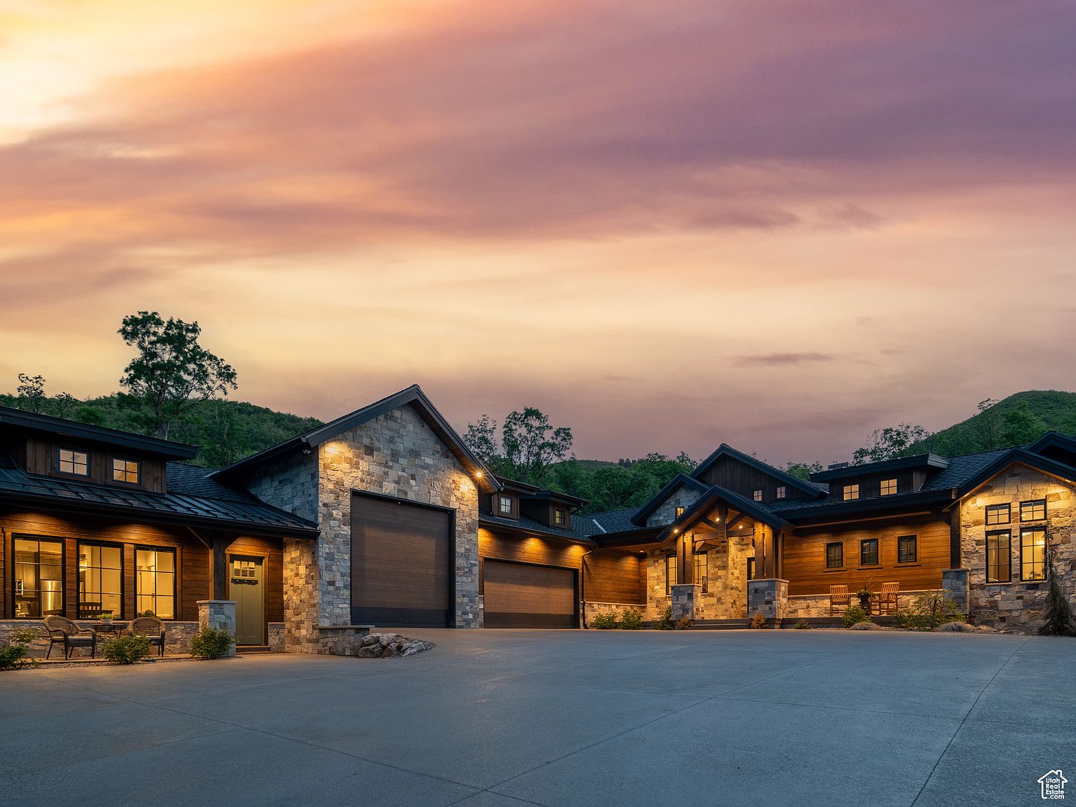 This is a front exterior view of a luxurious, sprawling home at dusk. The house features a combination of stone and wood siding, multiple garage doors, and well-lit windows that create a warm and inviting ambiance. The large concrete driveway provides ample parking space, and the surrounding landscape includes mature trees and a hillside backdrop.