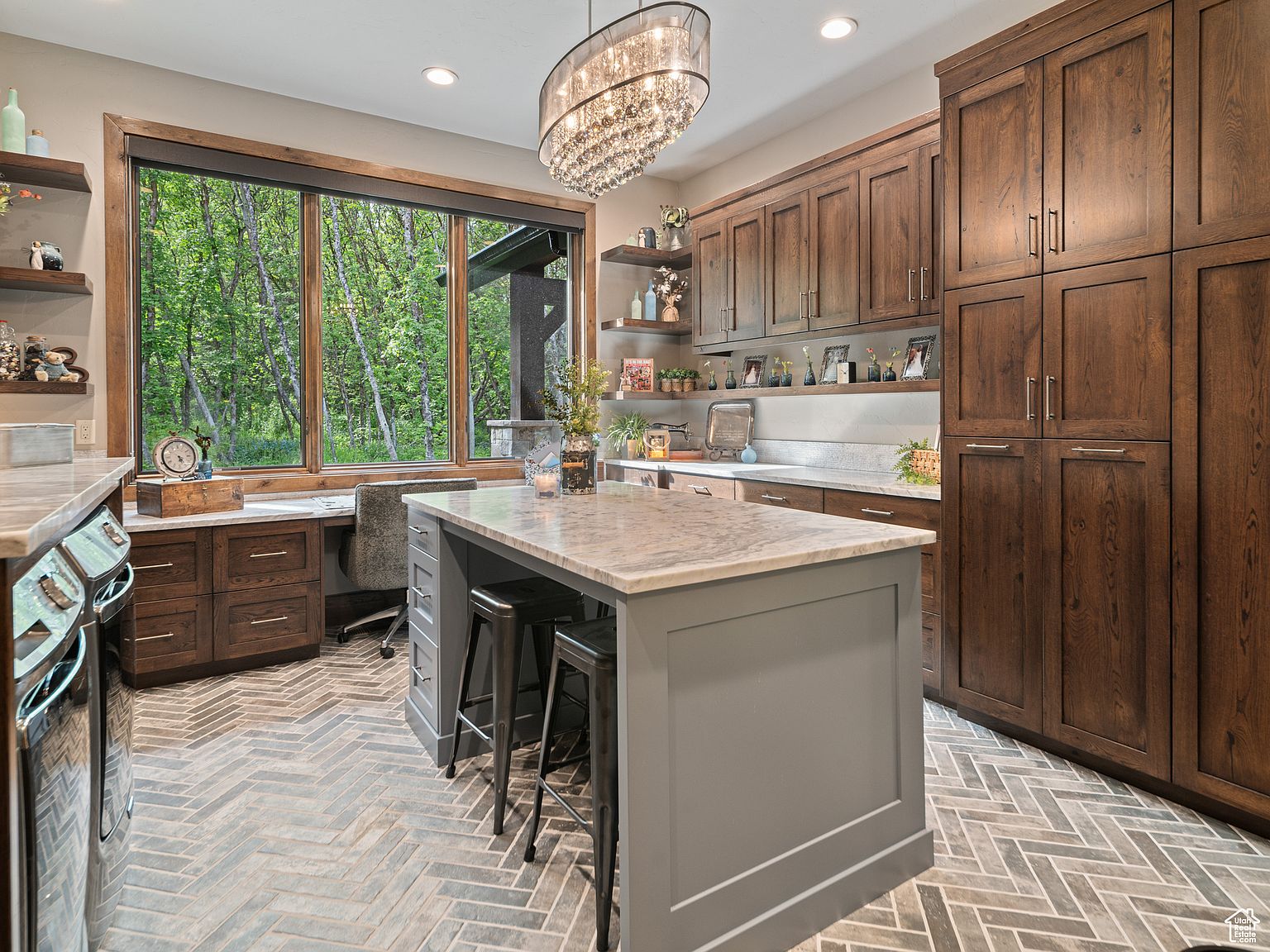 This interior shot showcases a well-appointed kitchen featuring dark wood cabinetry and a contrasting gray island with a marble countertop. A large window provides a view of a lush green forest, enhancing the natural light and creating a serene atmosphere. The herringbone-patterned tile floor adds a touch of elegance and sophistication to the space.