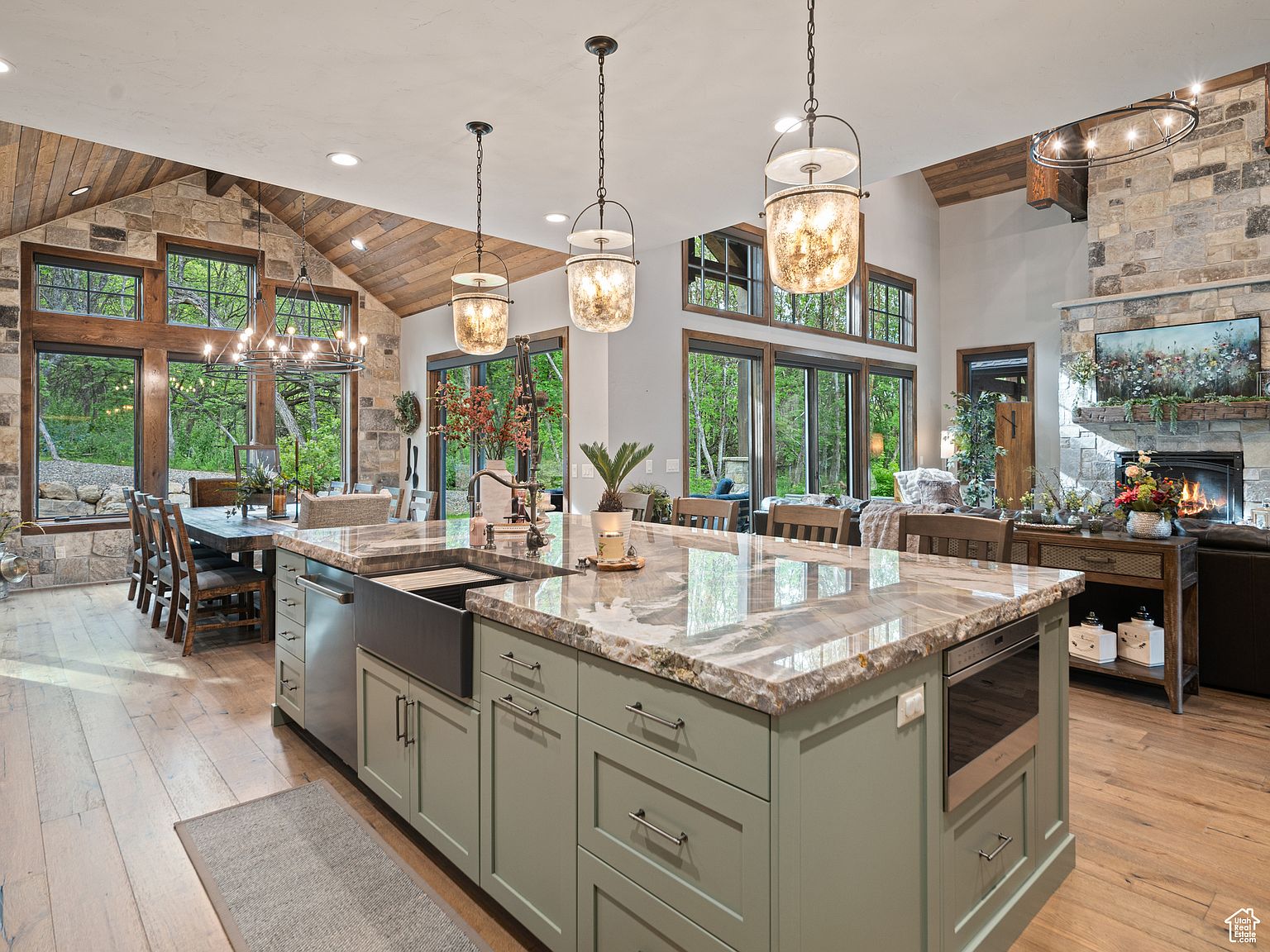 This is a spacious, open-concept kitchen and dining area featuring a large kitchen island with a farmhouse sink and granite countertops. The kitchen boasts custom cabinetry in a muted green hue, stainless steel appliances, and pendant lighting. The dining area is adjacent to the kitchen, with a large wooden table and chairs, and large windows offering views of the surrounding greenery. The overall style is rustic and luxurious.