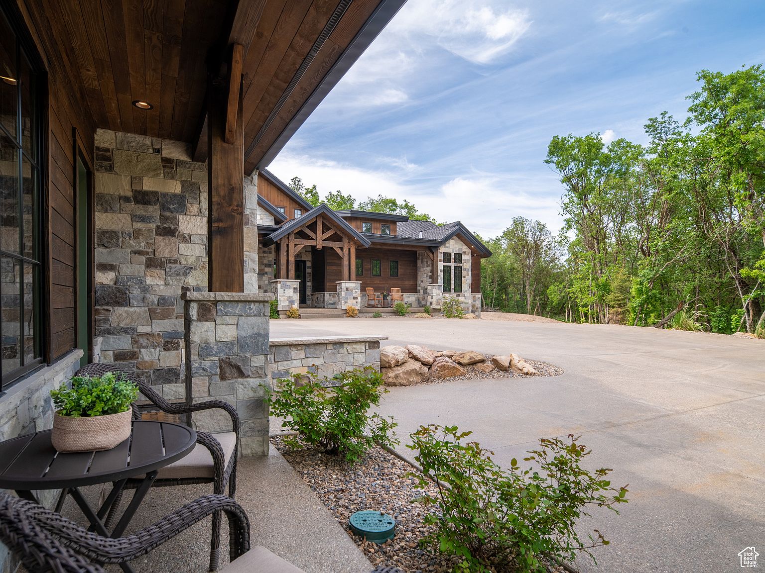 This is a front exterior view of a beautiful, modern home with a rustic touch. The house features a combination of stone and wood siding, creating a warm and inviting aesthetic. A spacious concrete driveway leads up to the house, and landscaping with rocks and greenery adds to the curb appeal. The overall impression is one of luxury and comfort.